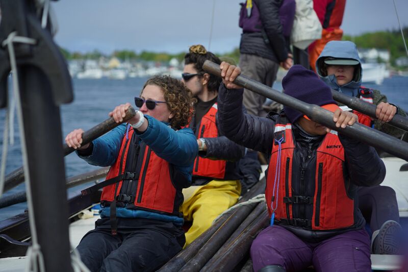 The image shows a group of people rowing a boat. They are wearing life jackets and appear to be on a body of water. The people are focused on rowing, and the boat seems to be moving forward. The background shows a shoreline with buildings and other boats.
